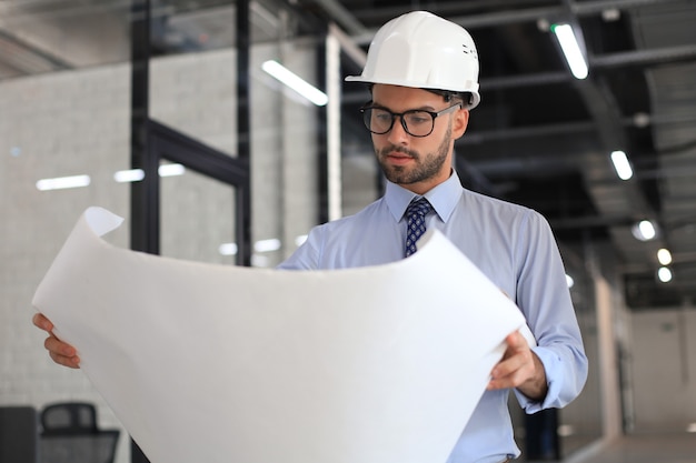 Arquitecto joven confiado en casco sosteniendo un plano y mirándolo.