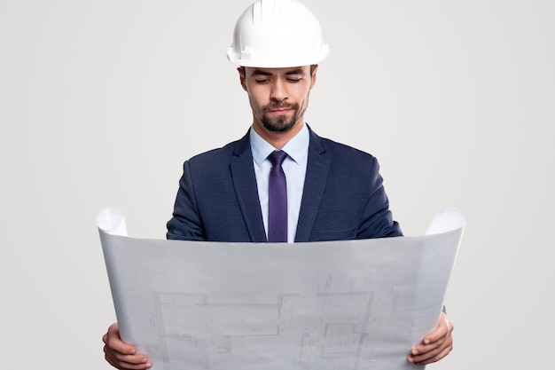 Arquitecto masculino barbudo atento en traje formal y casco de lectura plano con plan de construcción contra el fondo blanco.