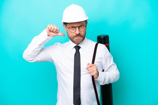 Arquitecto de mediana edad con casco y sosteniendo planos aislados de fondo azul mostrando el pulgar hacia abajo con expresión negativa