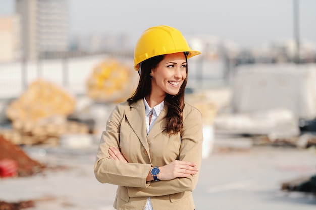 Arquitecto mujer atractiva con cabello castaño, sonrisa dentuda, casco en la cabeza y vestido elegante casual de pie en el sitio de construcción con los brazos cruzados.