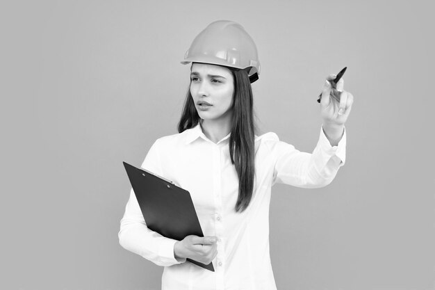 Arquitecto mujer en casco apuntando Mujer joven en casco mantenga portapapeles sobre fondo gris