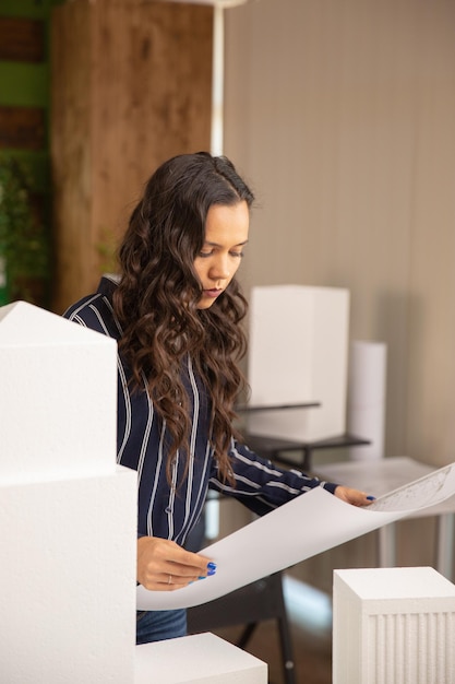 Arquitecto mujer caucásica mirando planos rodeados por modelos 3D de edificios