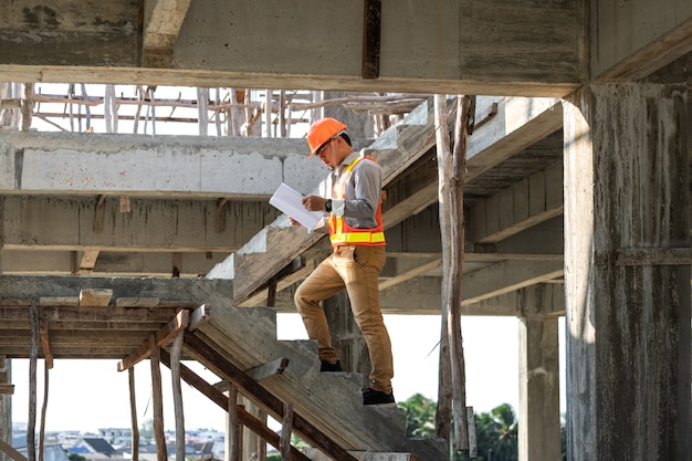 Foto arquitecto o ingeniero asiático sosteniendo buscando proyectos de planos y llevar casco en el sitio de construcción. él caminando por las escaleras.