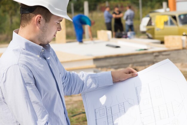 Arquitecto de pie estudiando un plano de mano en un sitio de construcción de una casa de nueva construcción