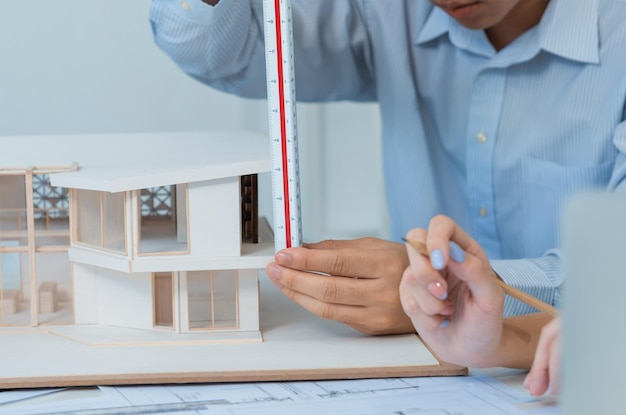 Foto arquitecto profesional asiático usando una regla para medir la longitud del modelo de casa mientras un joven y hermoso colega caucásico usa una computadora portátil para analizar datos en la mesa de reunión con el modelo de casa inmaculada