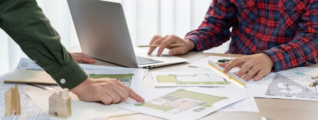 Arquitecto profesional diseña un plano usando una computadora portátil durante el gerente de proyecto muestra un punto de error en la mesa con un documento arquitectónico y bloques de madera dispersos alrededor de la Delineación de primer plano