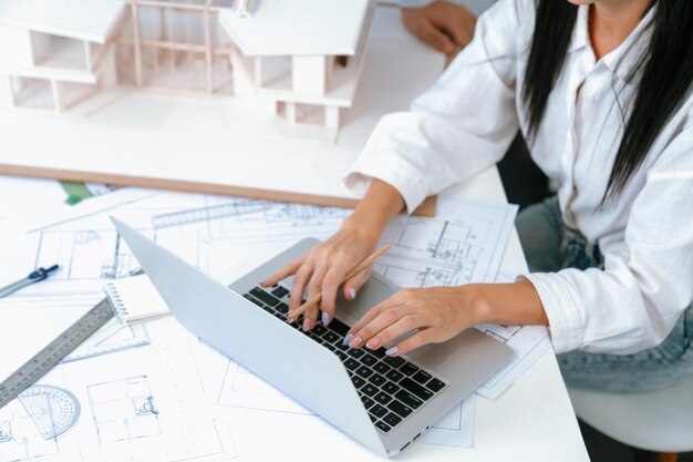 Arquitecto profesional masculino asiático usando una regla para medir la longitud del modelo de casa mientras un joven y hermoso colega caucásico usa una computadora portátil para analizar datos en la mesa de reuniones con el modelo de casa Inmaculada
