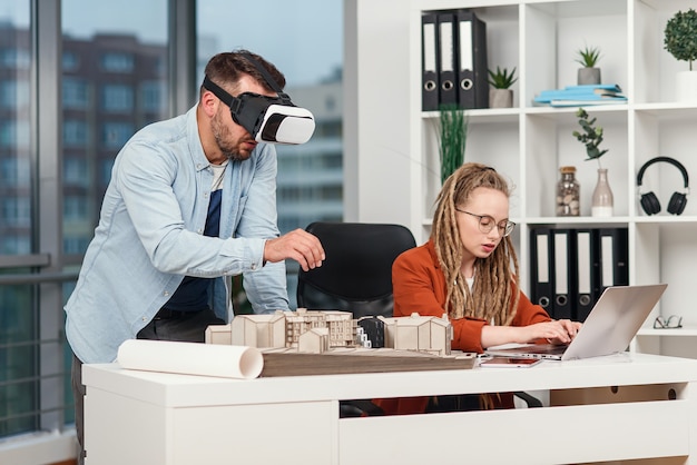 Arquitecto profesional masculino en gafas de realidad aumentada trabajando con maqueta de edificio y compañera de trabajo con laptop