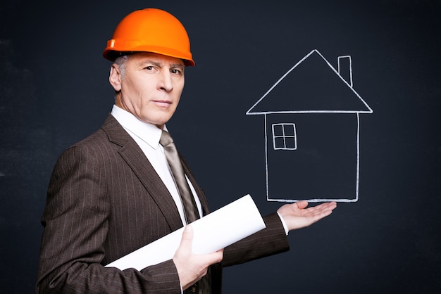 Arquitecto seguro. Hombre mayor seguro en ropa formal y casco sosteniendo una casa dibujada con tiza en su mano y llevando planos mientras está de pie contra la pizarra