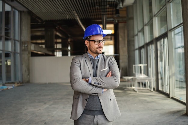 Arquitecto serio de pie en el edificio en proceso de construcción con los brazos cruzados.