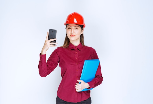 Arquitecto de sexo femenino en casco rojo con teléfono móvil y portapapeles.