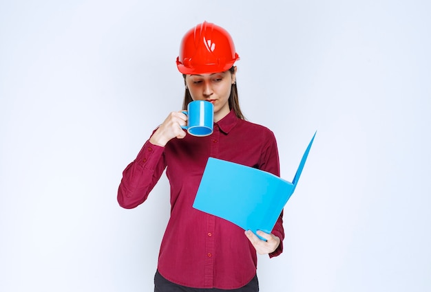 Arquitecto de sexo femenino en los documentos del casco rojo en la mano que tiene una pausa para el café.