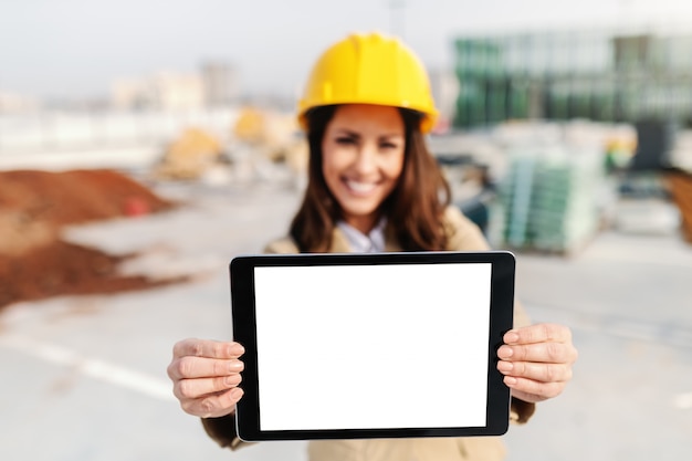 Arquitecto de sexo femenino hermoso sonriente con el casco en la tableta principal de la tenencia mientras que se coloca en el emplazamiento de la obra. Enfoque selectivo en tableta.