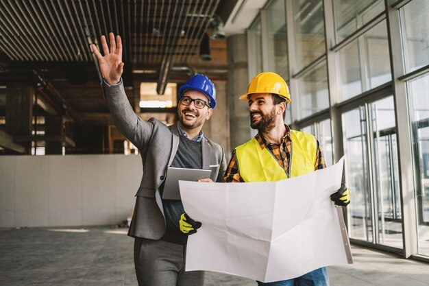 Arquitecto sonriente sosteniendo la tableta y mostrando al trabajador de la construcción algo que imaginó. Trabajador de la construcción con planos. Interior del sitio de construcción.