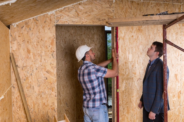 Arquitecto en traje chaqueta viendo trabajador de la construcción en casco tomando medidas del marco de la puerta con nivel interior nuevo hogar sin terminar