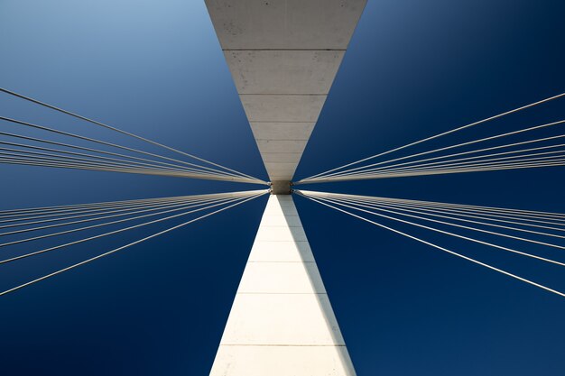 Arquitectura abstracta minimalista tomada con un pilar de hormigón blanco de un puente colgante con dos manojos de cables de suspensión contra un cielo azul claro