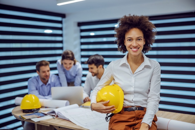 Foto arquiteta de raça mista sorridente, sentada na mesa do escritório e segurando o capacete