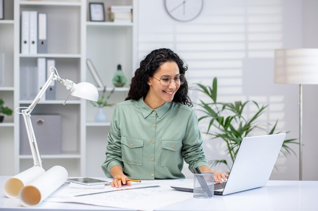 Arquiteta hispânica trabalhando em um laptop em um escritório moderno cercado de plantas e escritório