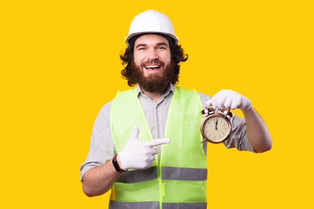 Arquiteto barbudo surpreso usando capacete e apontando para o despertador