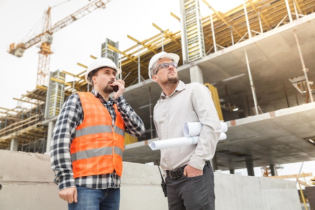 Arquiteto com plantas e desenvolvedor e um colete de segurança e walkie talkie no canteiro de obras