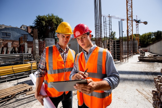 Arquiteto e engenheiro estrutural vestido com coletes e capacetes laranja discutem um projeto de construção no tablet no canteiro de obras ao ar livre com material de construção. .