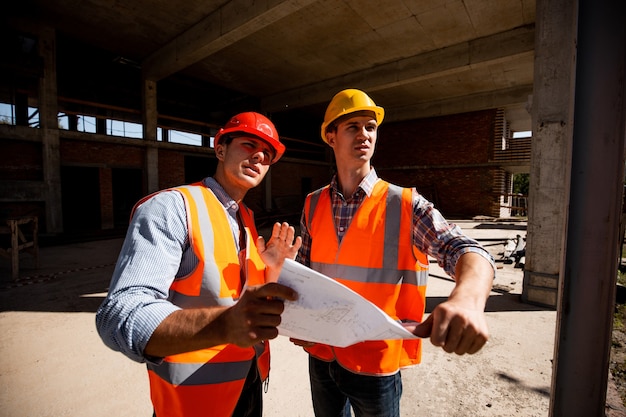 Arquiteto e engenheiro estrutural vestido com coletes laranja e capacetes, documentação de trabalho dentro do prédio em construção.