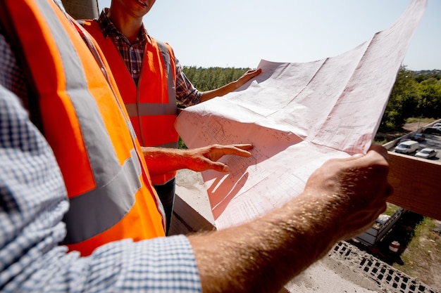 Arquiteto e engenheiro estrutural vestindo camisetas, coletes de trabalho laranja e capacetes exploram a documentação da construção no canteiro de obras dentro do prédio em construção.