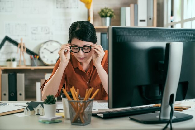Arquiteto jovem mulher asiática tirando os óculos no escritório. engenheira de construção feminina terminando projeto de desenho em plantas. designer de senhora colocando os cotovelos na mesa com papéis no computador desktop.