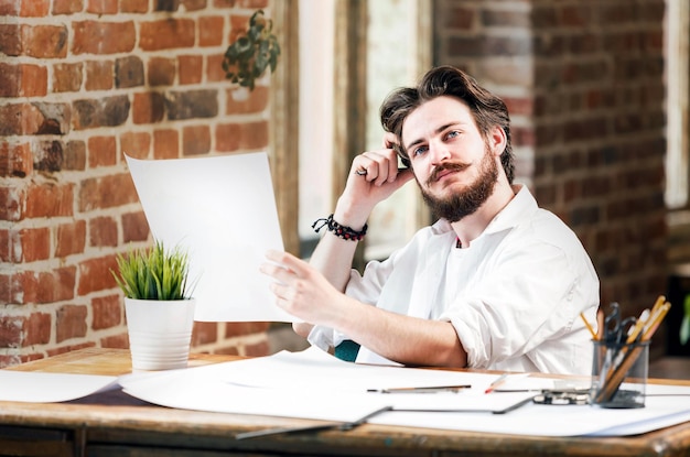 Arquiteto masculino hipster barbudo na camisa branca com divisores olhando a planta no escritório loft