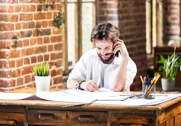 Arquiteto masculino incrível barbudo com lápis e planta usando telefone celular no escritório loft