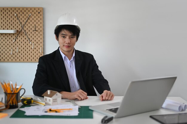 Arquiteto masculino profissional com capacete sentado na frente do laptop no escritório e olhando com confiança para uma câmera