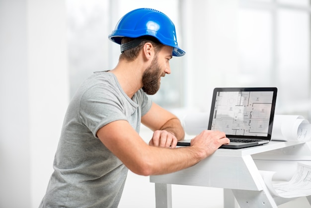 Foto arquiteto ou capataz bonito no capacete trabalhando com o laptop nos desenhos do apartamento no interior branco da estrutura