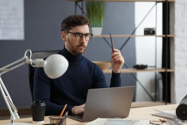 Arquiteto que trabalha no escritório com o laptop. O engenheiro pensa no plano arquitetônico, em busca de novas idéias para o projeto de construção. Retrato de homem barbudo bonito sentado no local de trabalho. Conceito de negócios.