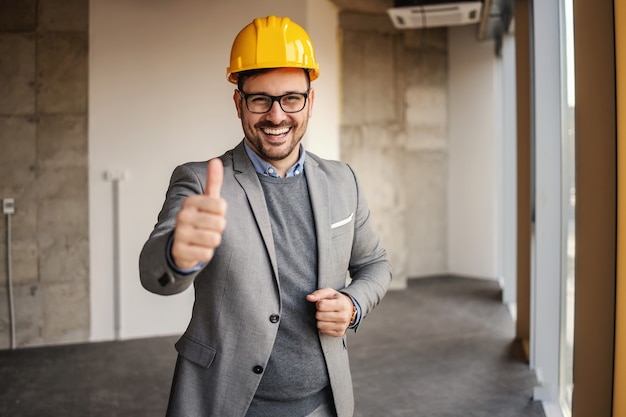 Arquiteto sorridente em pé no edifício em processo de construção e mostrando os polegares.