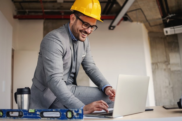 Arquiteto sorridente sentado na mesa no canteiro de obras e usando o laptop para melhorar o projeto