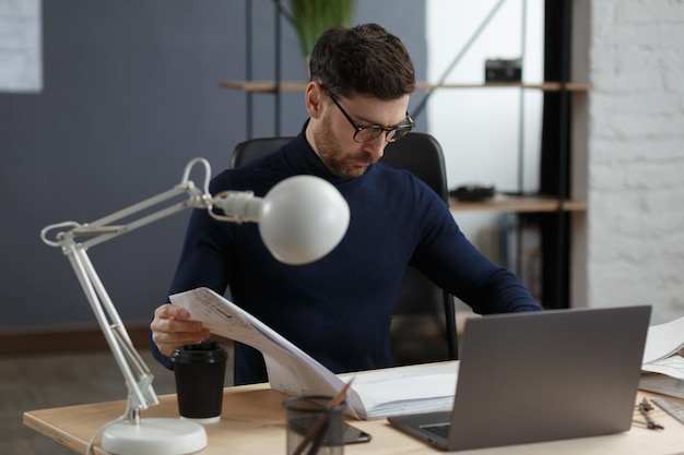 Arquiteto, trabalhando no escritório com plantas. Engenheiro, inspeciona o plano arquitetônico, esboçando um projeto de construção. Retrato de um homem barbudo bonito sentado no local de trabalho. Conceito de construção de negócios.