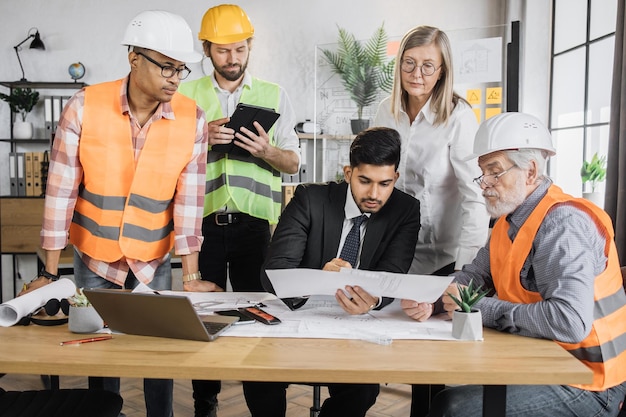 Arquitetos multirraciais em capacetes sentados à mesa com plantas e dispositivos modernos