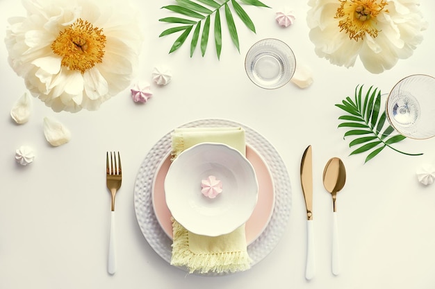 Arranjo de mesa de jantar de aniversário de verão flores de peônia amarelas pálidas e final da primavera postura plana de verão mesa de jantar branca utensílios brancos e dourados decorados com flores de peônia e folhas de palmeira exóticas