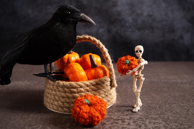 Arreglo decorativo de Halloween con una cesta de mimbre el esqueleto de un hombre que sostiene una calabaza y un cuervo negro sobre una mesa gris