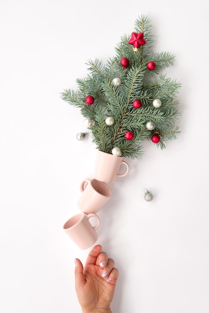 Arreglo de equilibrio creativo de Navidad. Las tazas de café sostienen la maceta con ramitas de árboles de Navidad decoradas con adornos navideños rojos y piñas. Mano sostiene la pirámide. Diseño navideño creativo.