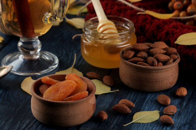 Arreglo de otoño con almendras, orejones, té, miel y una cálida bufanda y hojas amarillas sobre una mesa de madera azul oscuro.