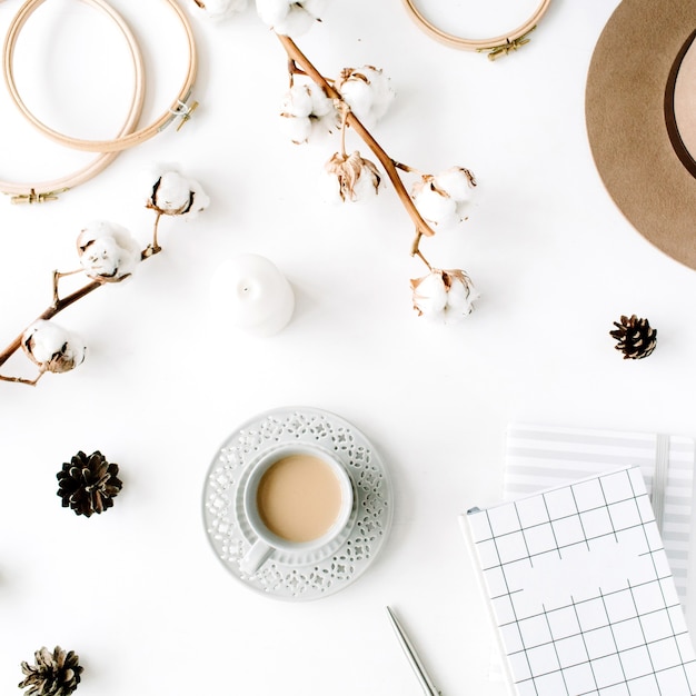 Arreglo plano de accesorios femeninos creativos de moda con café, rama de algodón y diario. Sombrero, rama de algodón, cuaderno, taza de café, cono de abeto, clips dorados sobre blanco