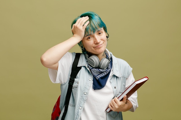 Arrependida jovem estudante usando fones de ouvido e bandana no pescoço e mochila segurando o caderno, mantendo a mão na cabeça, olhando para a câmera isolada no fundo verde azeitona