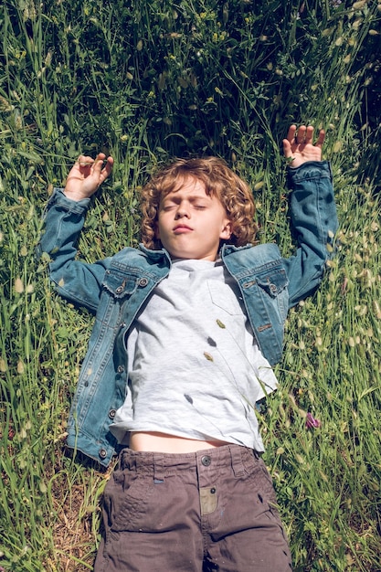 Desde arriba de un adorable niño preadolescente de pelo rizado durmiendo una siesta en la hierba verde del campo en un día soleado de verano