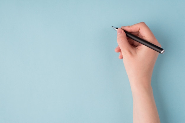 Arriba arriba arriba cerca de la vista en primera persona foto de la mano derecha de la niña sosteniendo un bolígrafo negro comenzando a escribir aislado sobre fondo azul pastel de color