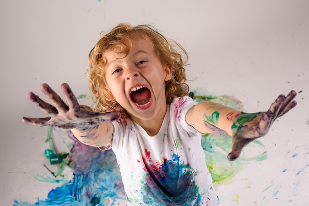 Desde arriba de un chico gracioso cubierto con pinturas coloridas mirando la cámara con cara emocionada sobre fondo blanco en un estudio desordenado