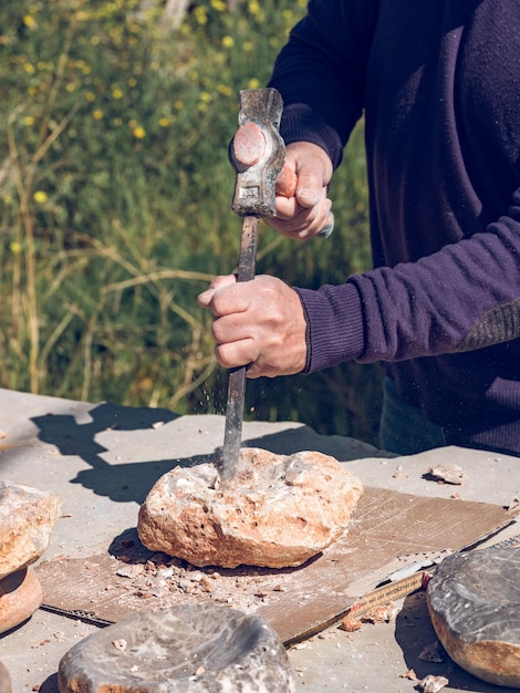 Desde arriba del cultivo, un artesano anciano irreconocible cortando roca sólida usando un martillo y un cincel mientras hace artesanías de piedra en un taller afuera