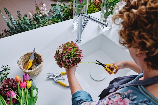 Desde arriba de una florista anónima arreglando un ramo de hortensias y cortando el tallo con tijeras cerca del fregadero en una tienda de flores