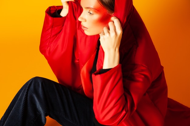 Desde arriba modelo de mujer solitaria con maquillaje creativo envuelto en un abrigo rojo de gran tamaño y mirando hacia otro lado contra el fondo amarillo