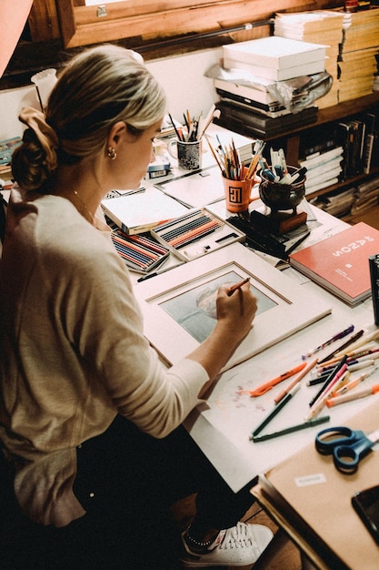 Desde arriba de una mujer joven concentrada creando un cuadro en la mesa con muchos suministros de arte Foto
